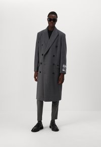 Cappotto grigio doppiopetto in un tessuto testurizzato, con grandi bottoni neri e un'etichetta sul maniche. Abbinato a pantaloni coordinati e scarpe nere.