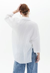 Camicia bianca in cotone strutturato con maniche a tre quarti e vestibilità ampia, caratterizzata da uno spacco sul retro per un tocco in più, abbinata a jeans blu.