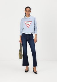 Felpa azzurro chiaro con logo triangolare rosso, indossata sopra una camicia a righe blu e bianche; abbinata a jeans svasati blu scuro e tacchi neri.