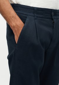 Mężczyzna noszący granatowe spodnie z ręką w bocznej kieszeni, elastycznym pasem oraz częściowo widoczną białą górą.