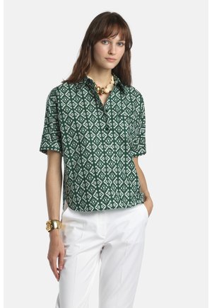 Femme portant une chemise à manches courtes à motif vert, un pantalon blanc, un bracelet en or massif et un grand collier de perles, debout avec une main dans la poche.