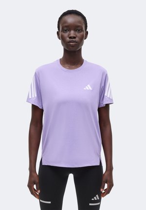 Persona que lleva una camiseta lila de Adidas con rayas blancas en las mangas y leggings negros con logotipos reflectantes de Adidas en los muslos, de pie inmóvil.
