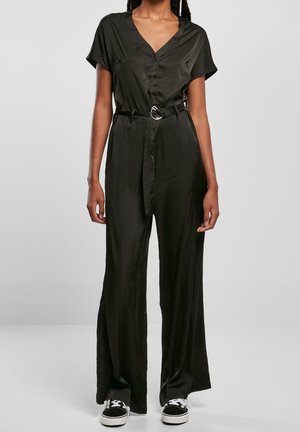 Woman wearing a black belted jumpsuit with short sleeves and wide legs paired with black and white sneakers.