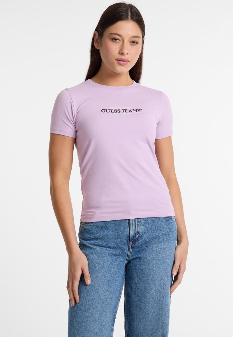 Violetinės figūrą aptempiančios marškinėlių su trumpomis rankovėmis ir apvalia iškirpte. Priekyje yra juodas „GUESS JEANS“ logotipas.