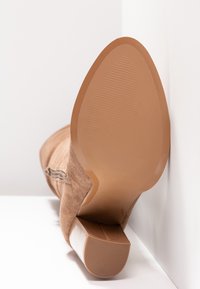 Stivale beige in suede con tacco e punta affusolata, rivolto verso la fotocamera, appoggiato su una superficie bianca in un angolo.