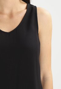Frau in einem ärmellosen schwarzen V-Ausschnitt-Shirt, das ihre rechte Schulter und den Oberarm vor einem hellen Hintergrund zeigt.