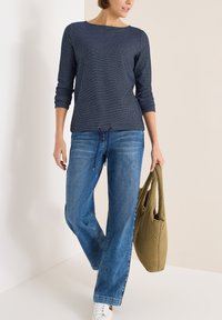 Marineblaues Langarmoberteil mit einem strukturierten Muster und Kordelzugdetails, kombiniert mit weit geschnittenen blauen Denim-Jeans und einer grünen Tasche.