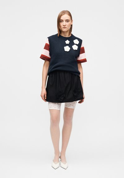 Marineblaues, gestricktes Gilet mit weißen Blumenapplikationen, gestreifte kurze Ärmel, schwarzer Rock mit floralen Akzenten, Spitzen-Shorts blitzen hervor und weiße Absätze.