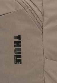 Bolso Thule en tejido de color marrón claro con un acabado texturizado; cuenta con logotipo negro y cremalleras. El diseño incluye múltiples bolsillos para almacenamiento.