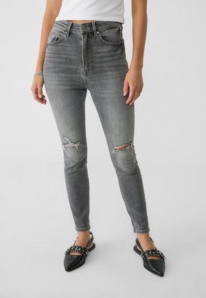 Vrouw die grijze, hoog getailleerde, gescheurde skinny jeans draagt en zwarte puntige platte schoenen met bandjes en zilveren studs, staand op een grijze vloer.