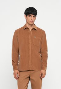 Brown corduroy shirt with long sleeves, button-up front, chest pocket, and embroidered green logo. Smooth texture and classic collar design.