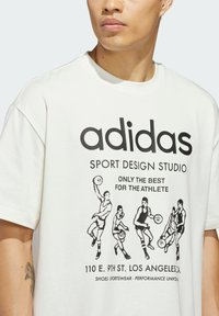 Biela bavlnená tričko s čiernym grafickým potlačou basketbalistov a textom, vrátane "adidas" a "SPORT DESIGN STUDIO."