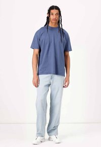 T-shirt de algodão azul com um corte descontraído e mangas curtas, combinado com jeans azul claro e ténis brancos. Design e estilo minimalistas.