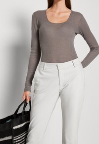 Kvinna som bär en figurnära grå långärmad topp och vita byxor, håller en svart-och-vit randig tote-väska med ena handen.