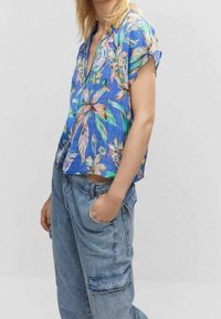 Top corto a maniche corte blu a fiori con scollatura a V, caratterizzato da motivi rosa, verdi e gialli; abbinato a jeans baggy azzurri chiari.