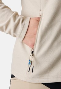 Chemise beige à manches longues avec une poche latérale zippée. Caractérisée par un tissu texturé, un cordon de serrage avec des accents noirs, et un petit emblème bleu et jaune.