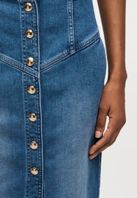 Κοντινή λήψη μπλε τζιν φούστας με χρυσά μεταλλικά κουμπιά κατά μήκος του εμπρός μέρους, με λεπτομέρειες ραφής και το χέρι ενός ατόμου που ακουμπάει δίπλα του.