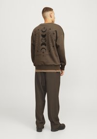 Sudadera marrón con un gráfico de la luna y el sol en la parte posterior. Corte holgado con puños y dobladillo acanalados. Llevada con pantalones marrones a juego.