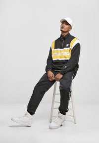Jachetă de vânt neagră și galbenă cu dungi albe, având guler cu fermoar și logo. Asortată cu pantaloni sport negri și adidași albi cu glezna înaltă.