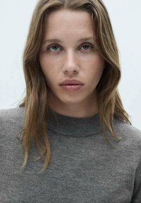 Siv trikotan pulover z visokim ovratnikom, ki se ponaša z mehkimi materiali in minimalističnim dizajnom. Model ima srednje dolge valovite lase in nevtralen make-up.