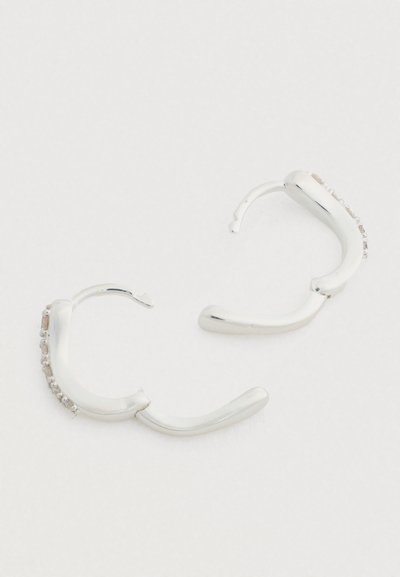 Cercei hoop din argint cu un design ondulat, având cristale clare de-a lungul unei părți și o textură netedă, lustruită.