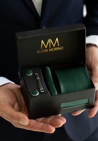 Zaļā kaklasaite, saskaņoti aproču pogas un kaklasaite klips, kas iesaiņoti melnā kastē ar zelta logo "MASSI MORINO". Redzami teksturēti materiāli.