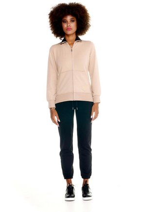 Une femme porte une veste beige zippée, un pantalon de jogging noir et des baskets noires sur fond blanc.
