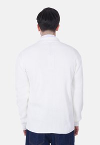 Maglione bianco lavorato a maglia con un motivo testurizzato, dotato di colletto e maniche lunghe. La vista posteriore mostra un taglio dritto con un orlo leggermente più lungo.