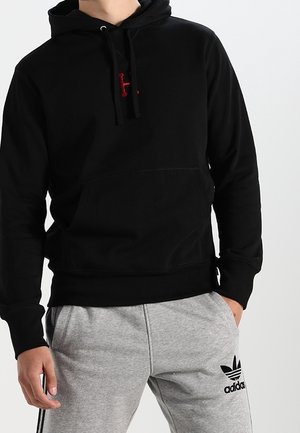 Osoba ubrana w czarną bluzę z kapturem z czerwonym logo i jasno szare spodnie dresowe Adidas z czarnym logo trefoil i bocznymi paskami, ręce w kieszeniach.