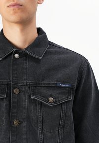Melnas džinsu jaka ar apkakli, divām krūšu kabatām, zelta tonim aparatūru un mazu zilu birku ar uzrakstu "Pepe Jeans London."