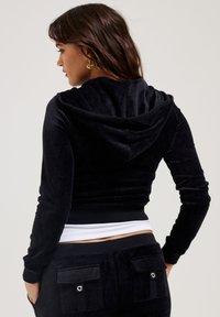 Black cropped velvet hoodie with a hood, long sleeves, and a contrasting white waistband. Features a smooth texture and fitted design.