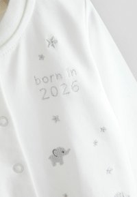 Body alb pentru bebeluși, cu text brodat argintiu „născut în 2026” și modele mici de elefant și stele. Material moale și închideri cu capse.