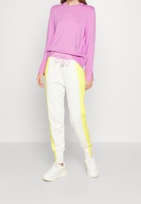 Maglietta viola a maniche lunghe con aperture laterali, abbinata a pantaloni jogger bianchi con strisce gialle ai lati. Sneakers bianche completano l'outfit.