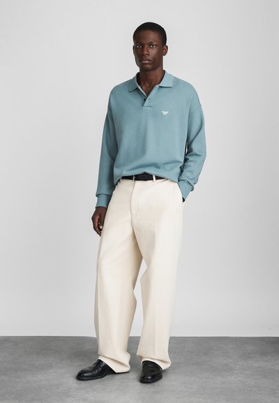 Ljusblå långärmad polo med krage och brodderad bröstlogga, parat med vita vida byxor och svarta skor.