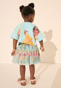Un tricou din bumbac de culoare teal are grafice cu prințese Disney pe spate, asortat cu o fustă transparentă, cu imprimeu, care îl reprezintă pe diverse prințese.
