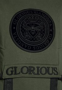 Rond zwart schild met een bij, kroon en decoratieve elementen geborduurd op donkergroene stof, met "GLORIOUS" eronder gestikt.