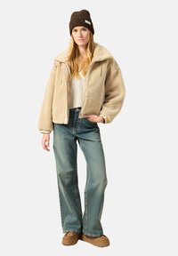 Chaqueta de felpa beige con corte oversized, cierre de cremallera y puños elásticos; combinada con unos vaqueros de pierna ancha azules y un gorro de punto marrón.