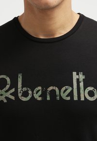 Černé tričko s krátkým rukávem s texturovanou zelenou logografií, která zobrazuje název značky "benetton" napříč hrudí. Kulatý výstřih.