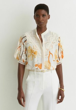 Žena oblečená v bielej blúzke s oranžovým kvetinovým vzorom a detailom z dierkovanej čipky, v kombinácii s bielymi nohavicami s vysokým pásom a béžovým opaskom.