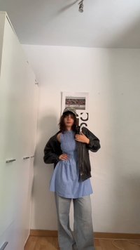Άτομο με μπλε φορεματάκι χωρίς μανίκια πάνω από φαρδιά τζιν, μαύρο δερμάτινο μπουφάν και καπέλο με σχέδιο, στέκεται σε έναν αραιά επιπλωμένο χώρο.