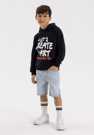 Sweat à capuche noir avec le texte graphique "Créons de l'art" en blanc et orange, associé à un short en denim bleu clair et des baskets blanches avec des rayures noires.