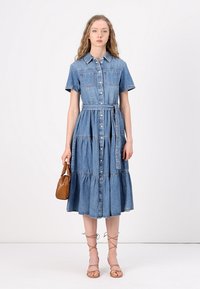 Robe chemise en denim à manches courtes, avec des boutons sur le devant, deux poches poitrine et une taille ceinturée. Jupe plissée fluide, de couleur bleue.