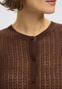 Brun strikket cardigan med et teksturert mønster, med rund hals og tre brune knapper foran.