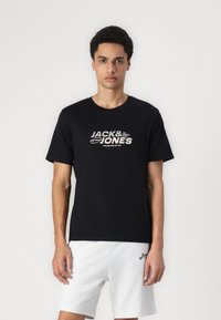 Svart bomull T-skjorte med korte ermer, med hvit tekst "JACK & JONES" i en fet grafisk stil. Kombinert med grå shorts.