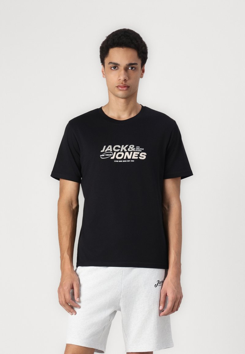 Svart bomull T-skjorte med korte ermer, med hvit tekst "JACK & JONES" i en fet grafisk stil. Kombinert med grå shorts.