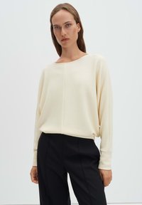 Frau mit glattem Haar trägt einen cremefarbenen langärmligen Strickpullover und schwarze maßgeschneiderte Hose vor einem schlichten hellen Hintergrund.