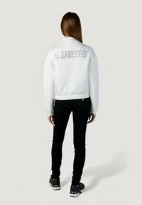 Chaqueta blanca con cuello alto, con un gran logo "GUESS" en pedrería en la parte trasera. Combinada con vaqueros pitillo negros y zapatillas.