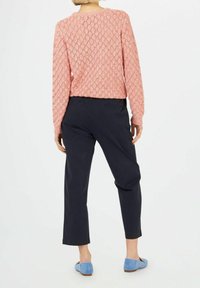 Frau trägt einen rosa Strickpullover, schwarze, kurze Hosen und helle blaue Ballerinas, steht mit dem Rücken zur Kamera vor einem weißen Hintergrund.