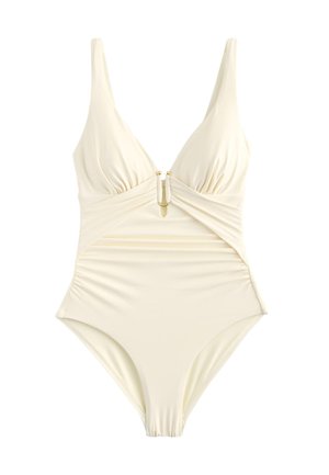 Kvinde med krøllet mørkt hår iført en beige ét-stykke badedragt står på en sandstrand med havets bølger i baggrunden og smiler med lukkede øjne.