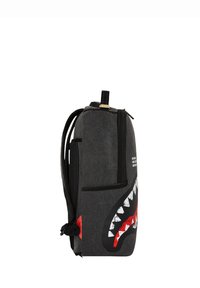 Mochila gris oscuro con correas negras, un bolsillo con cremallera en la parte frontal y un gráfico de boca de tiburón rojo y blanco en el lateral.